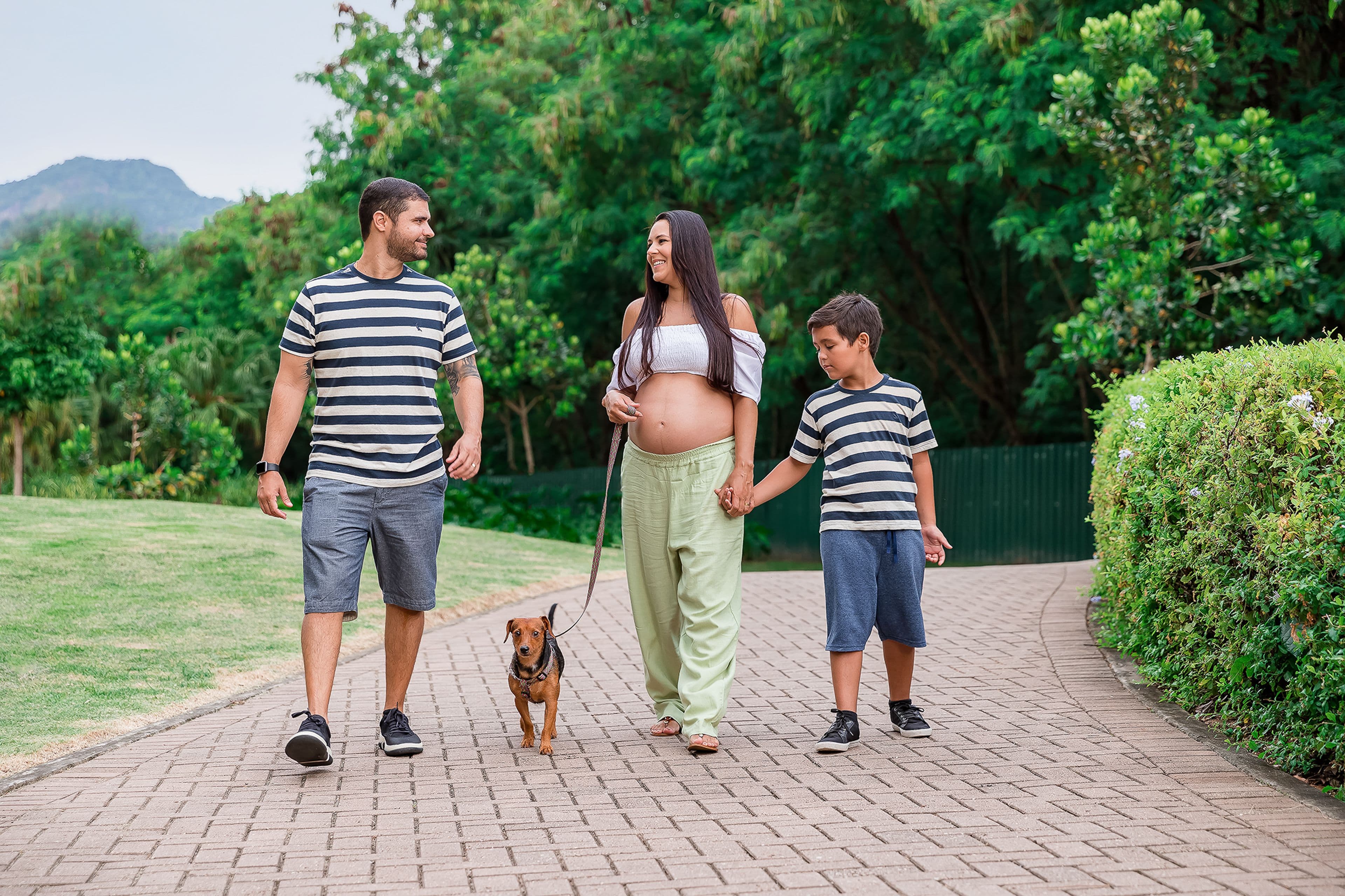 Gestante caminhando num parque de mãos dadas com o filho, ao lado do companheiro e do cachorro