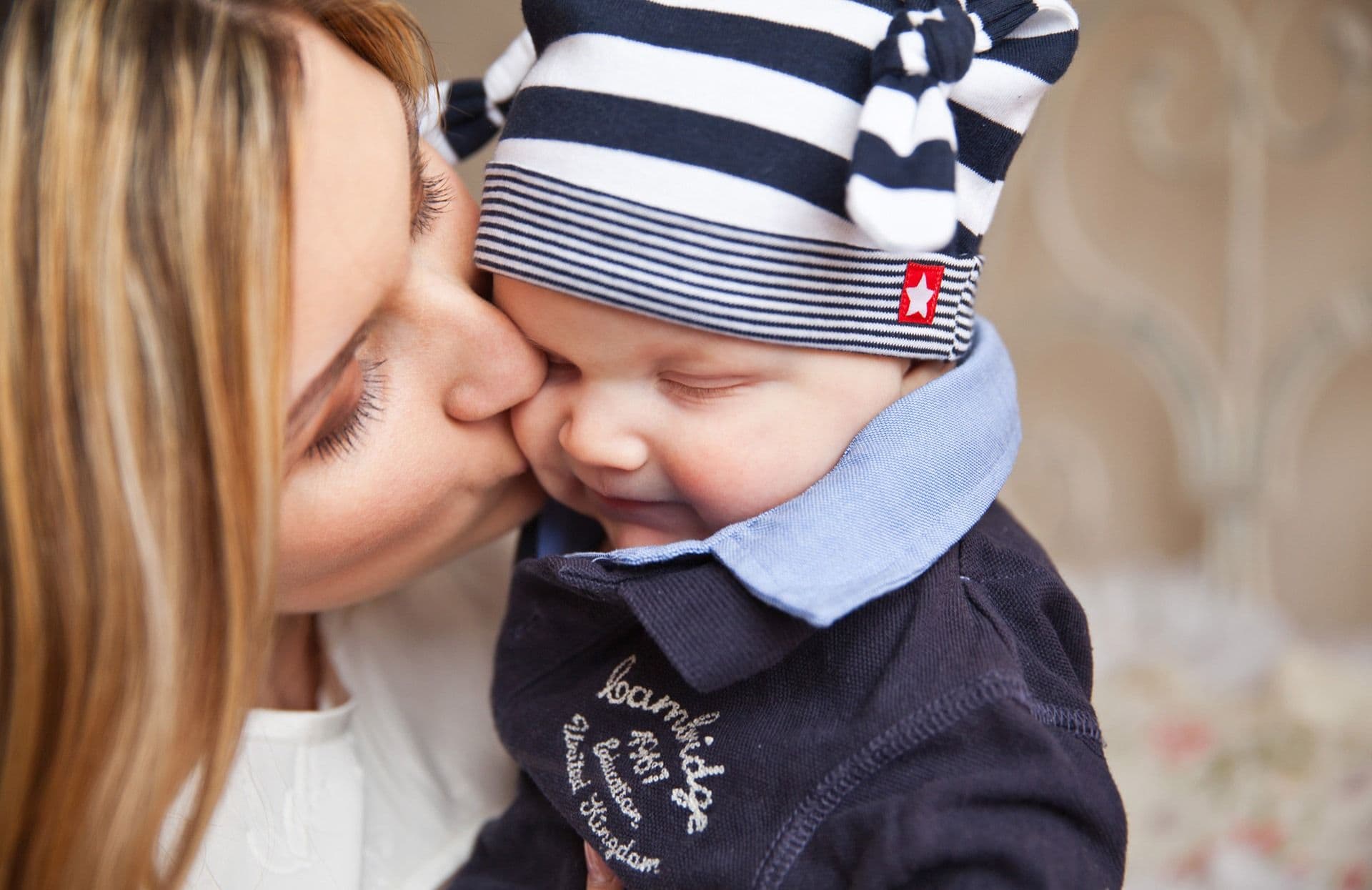 Mãe beijando a bochecha de um bebê de gorro listrado, que sorri de olhos fechados
