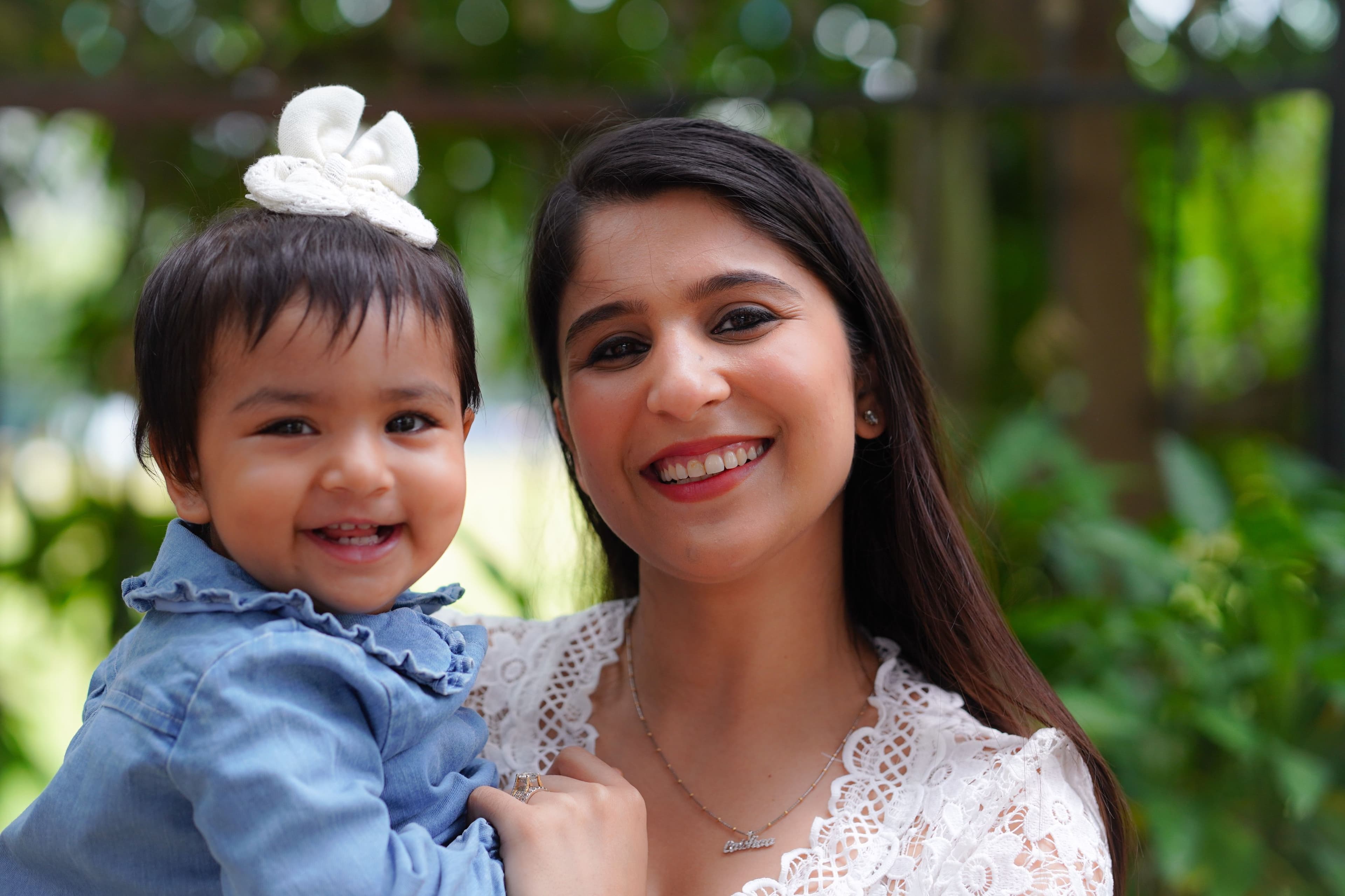 Mãe sorrindo com um bebê de cerca de 1 ano no colo, jardim ao fundo